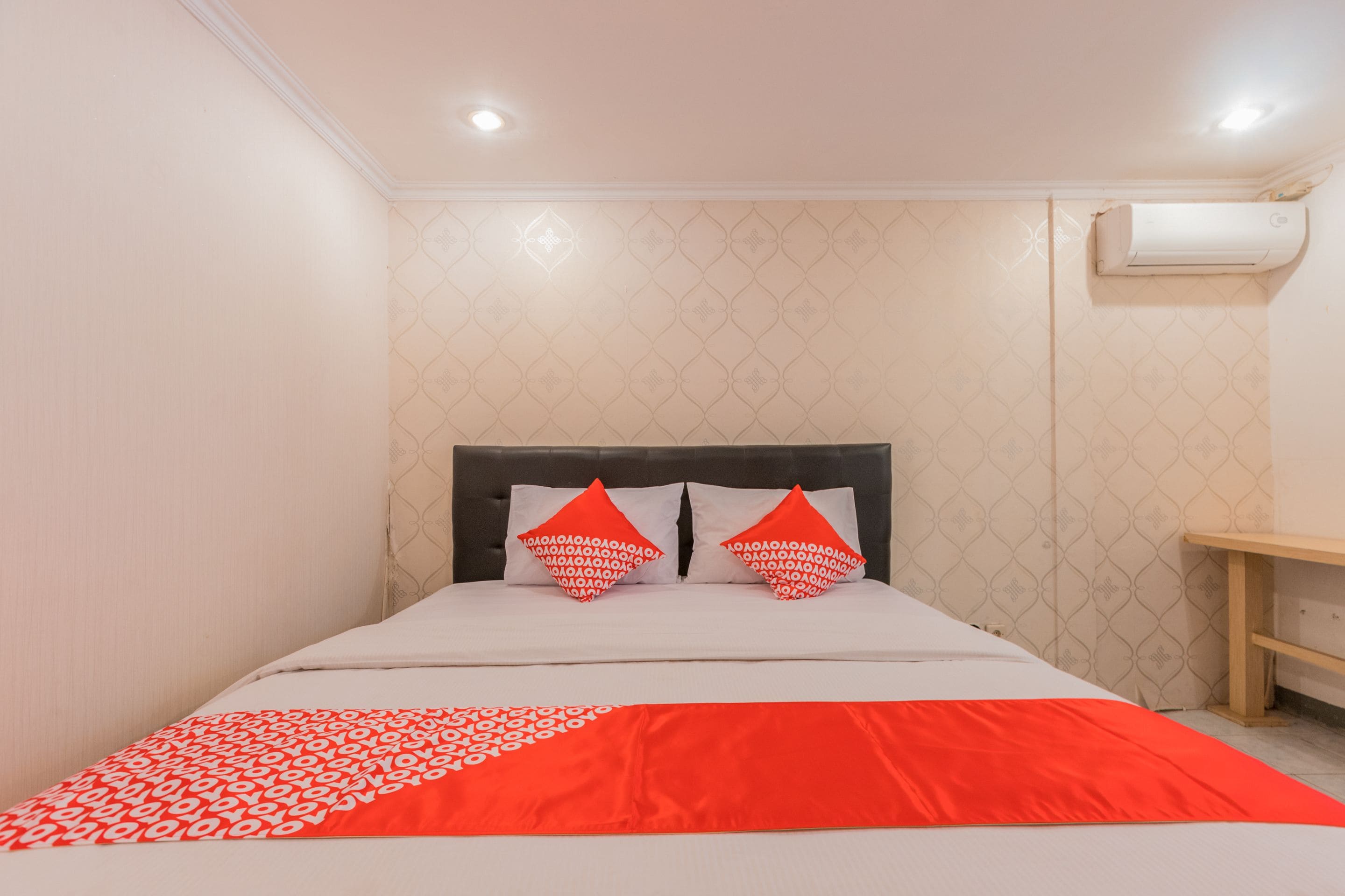 Una cama bien tendida con una colcha estampada, dos almohadas con un diseño geométrico y una mesita de noche con un cajón de color claro.