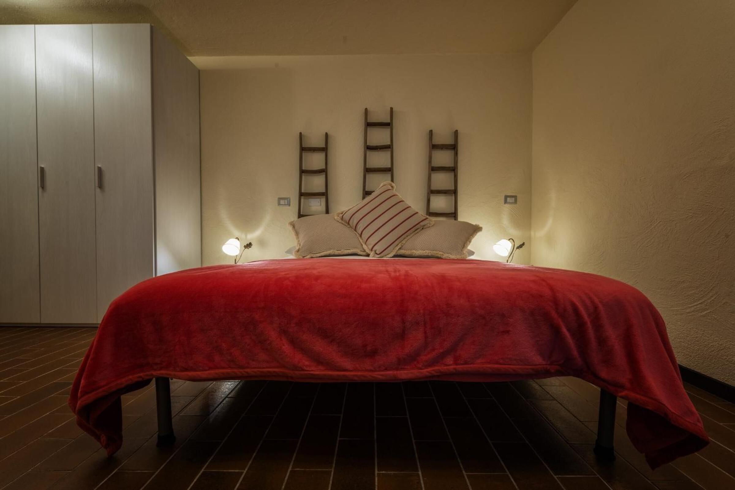 A bedroom with a bed, two bedside lamps, a wardrobe, and three wooden ladders against the wall.