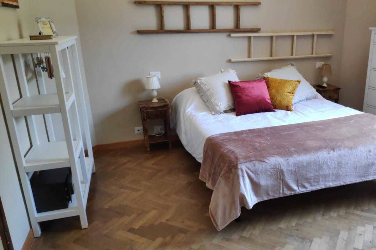 Un dormitorio con suelo de madera, una cama con manta rosa y dorada, un aparador blanco y una estantería de madera.