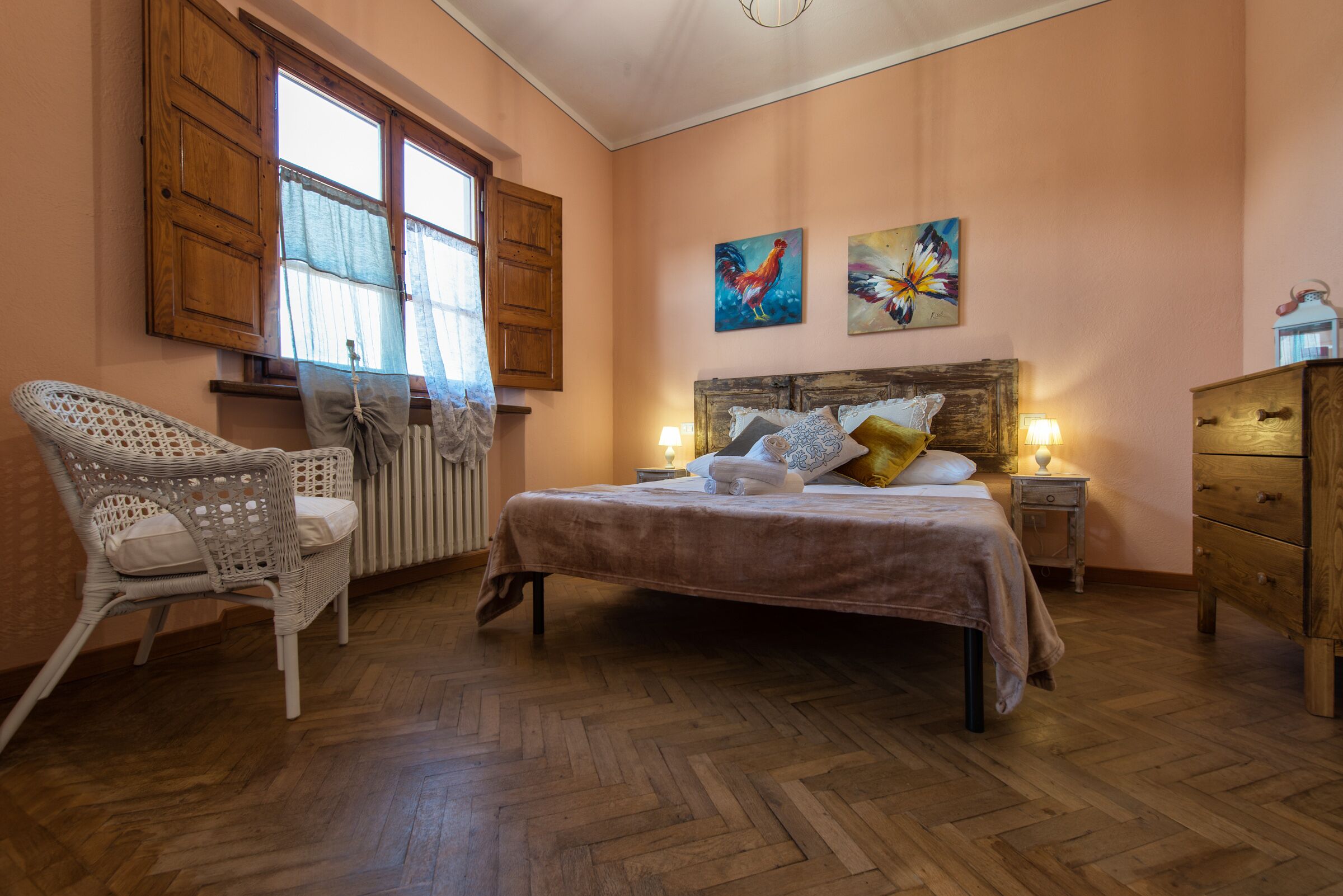 A bedroom with a wooden bed, a wicker chair, a wooden dresser, and two paintings on the wall.