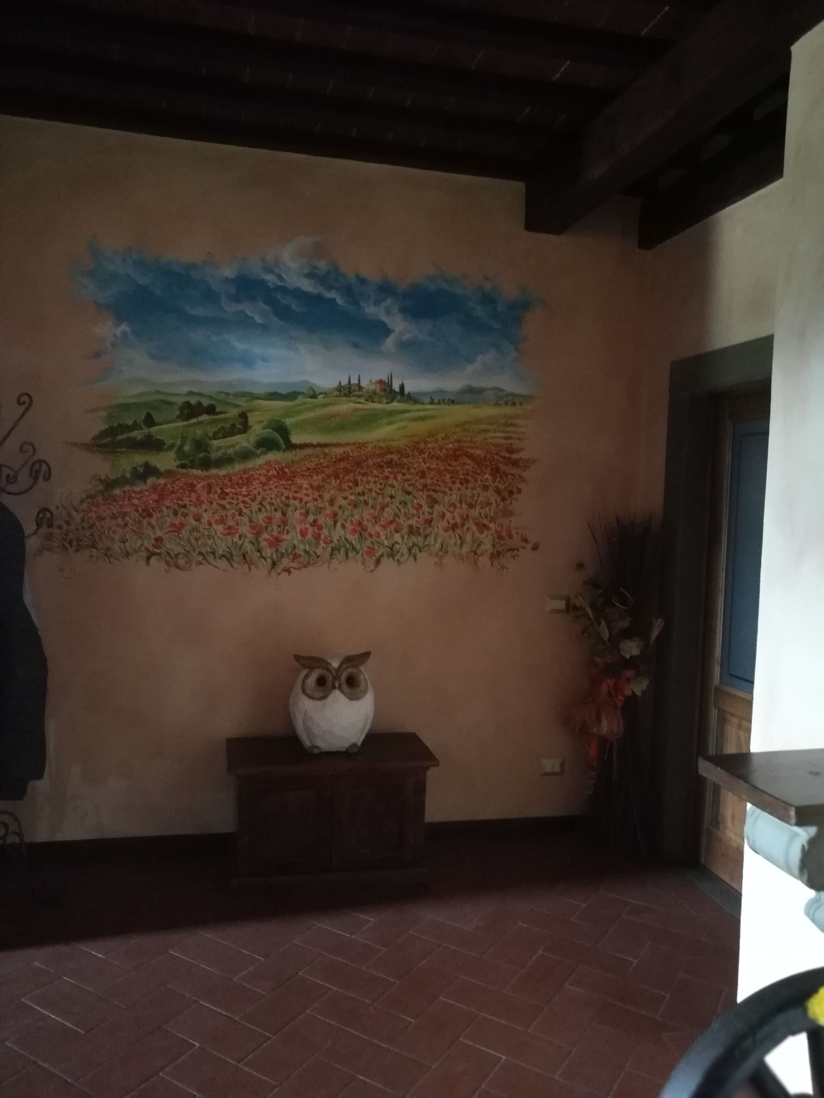 A room with a mural of a landscape, a wooden bench with an owl sculpture, and a brick floor.
