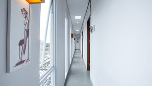 Un long couloir étroit aux murs blancs et au sol gris, avec une grande fenêtre sur le côté gauche et une porte sur le côté droit.