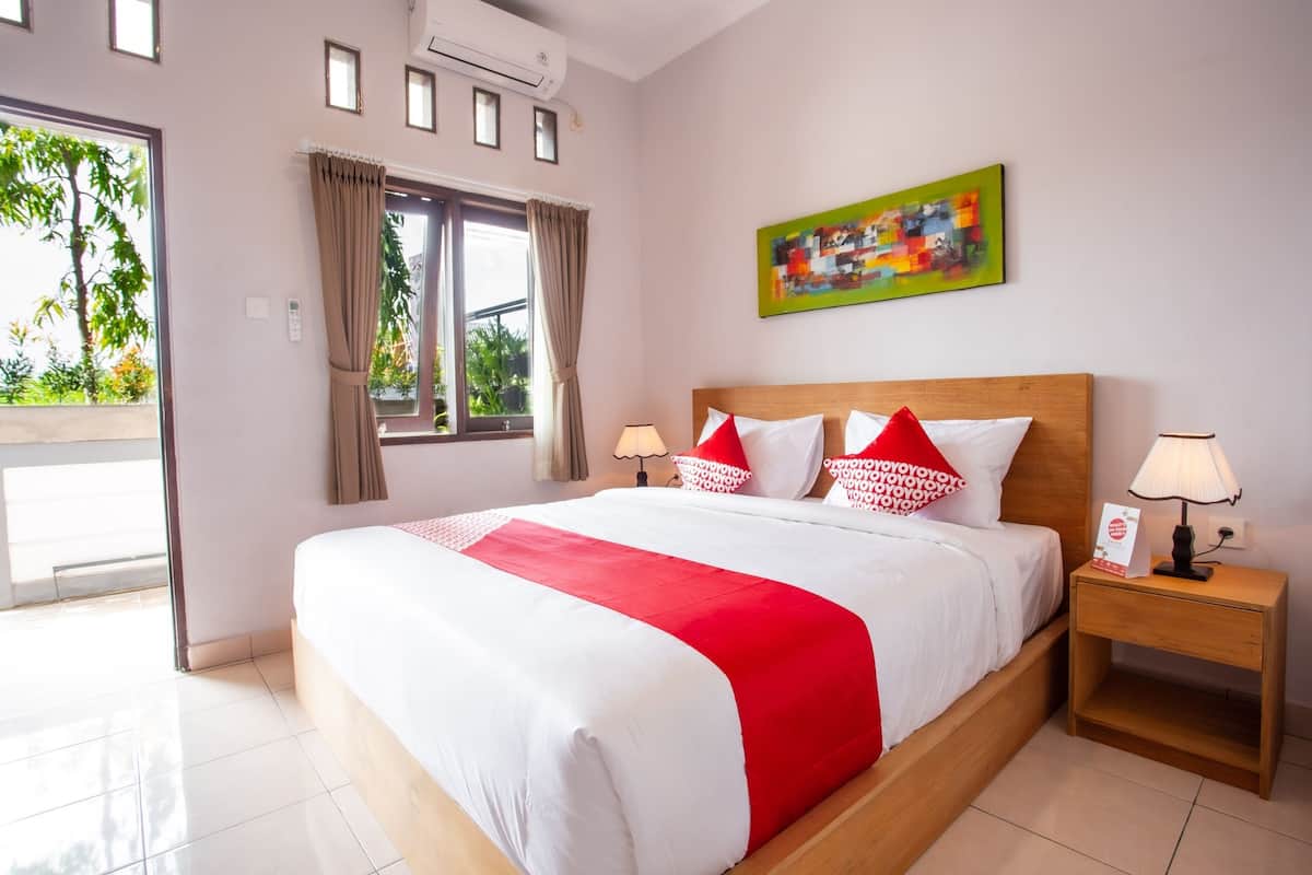 Habitación de hotel con una cama grande, cabecera de madera, dos mesitas de noche con lámparas, una ventana con cortinas y un cuadro abstracto de colores en la pared.