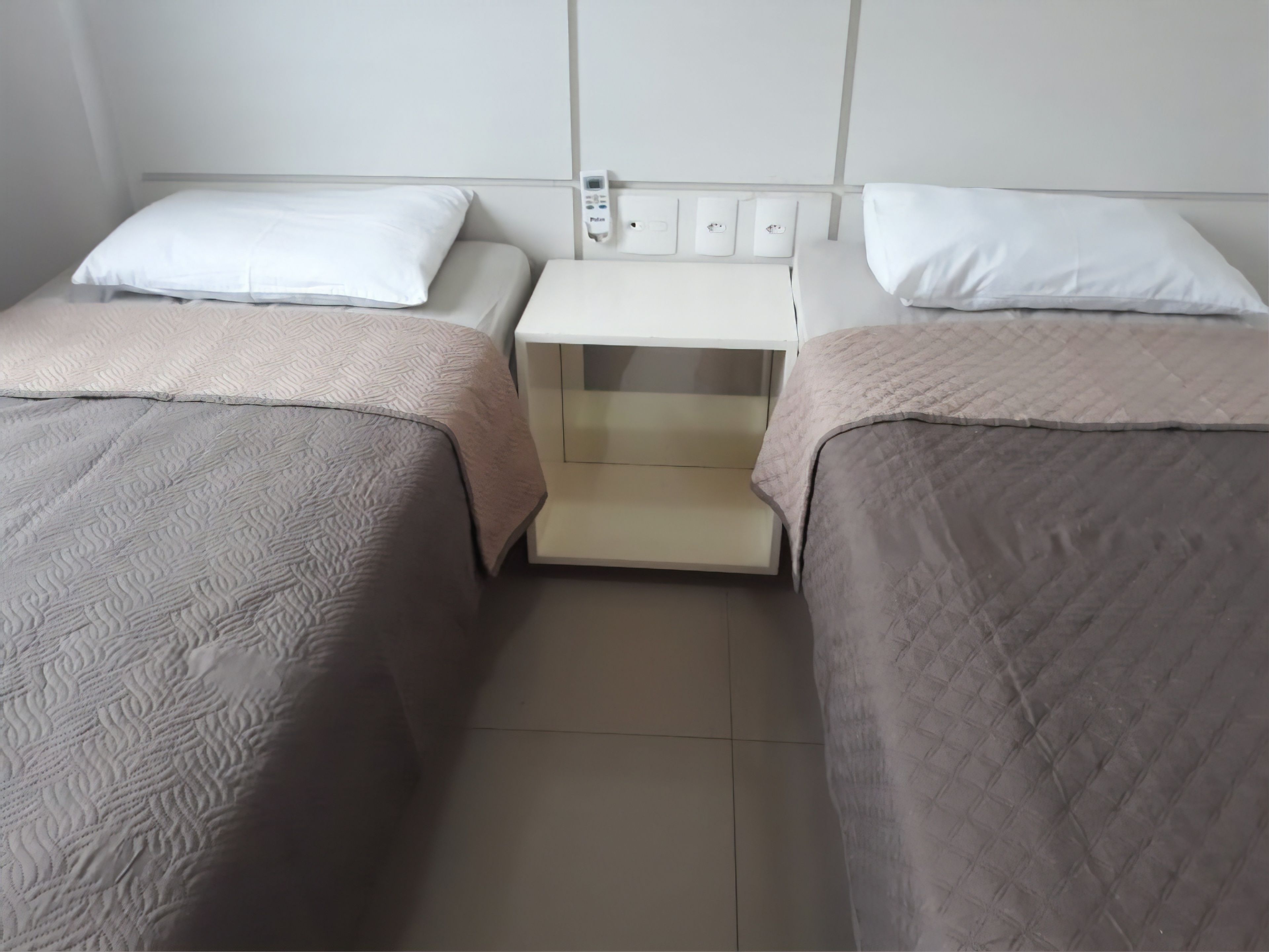 Ein Hotelzimmer mit zwei Betten, jeweils mit Kissen und einer gesteppten Decke. Ein kleiner Nachttisch mit Lampe und Steckdose ist vorhanden.