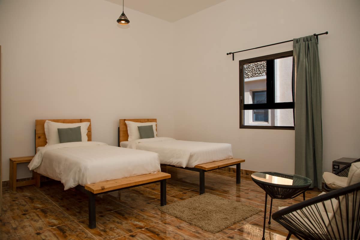A room with two single beds, a small table, a chair, and a window with curtains.