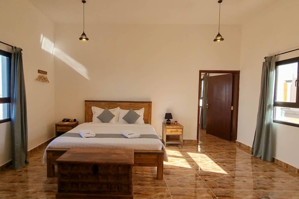 A bedroom with a wooden bed, two bedside tables, a nightstand, a bench, and a door leading to another room.