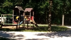 A playground with a slide and climbing structures, surrounded by trees and a dirt path. - Les Voiles de Laives (Laives)