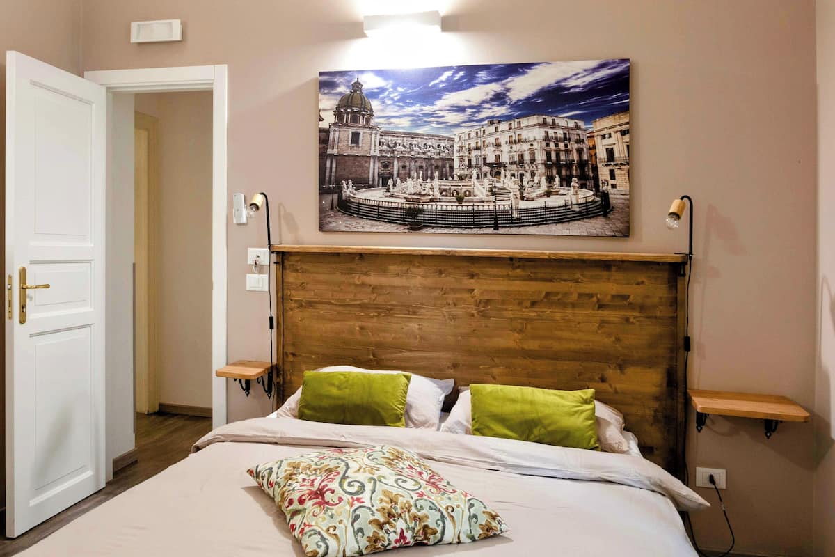 A bedroom with a wooden headboard, a bed with white bedding and green pillows, a wall-mounted picture of a cityscape, and bedside tables with lamps.