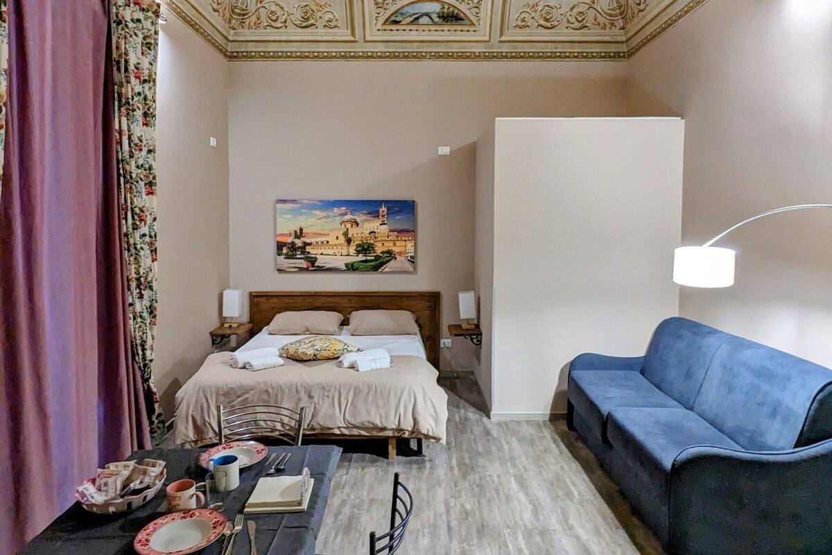 A hotel room with a bed, a dining table with chairs, a sofa, and a painting on the wall.