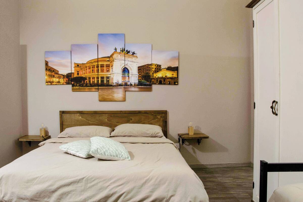 A bedroom with a large bed, a wooden headboard, two bedside tables with lamps, and a wall-mounted artwork depicting a building.