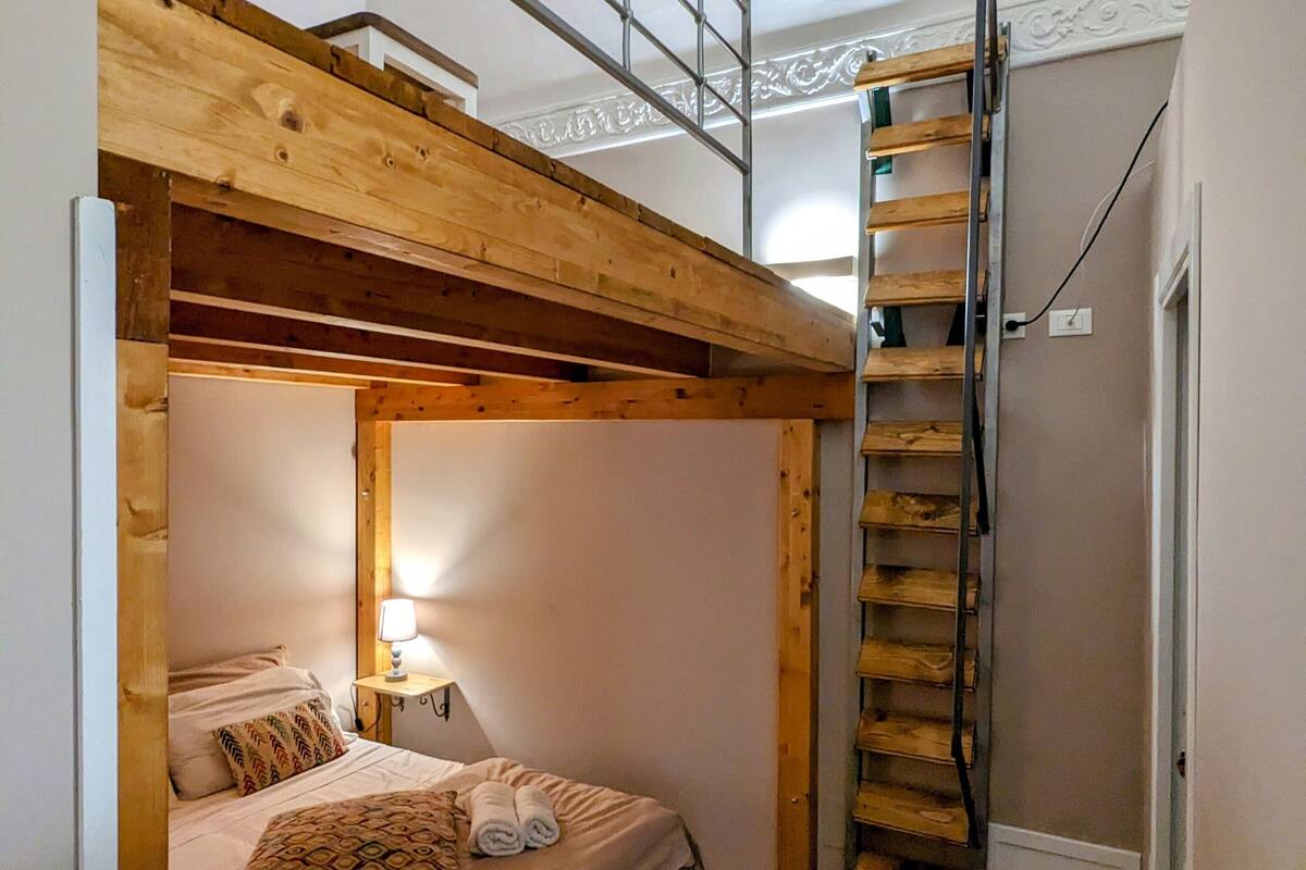 A room with a bunk bed, a ladder, a bedside table with a lamp, and a small window.