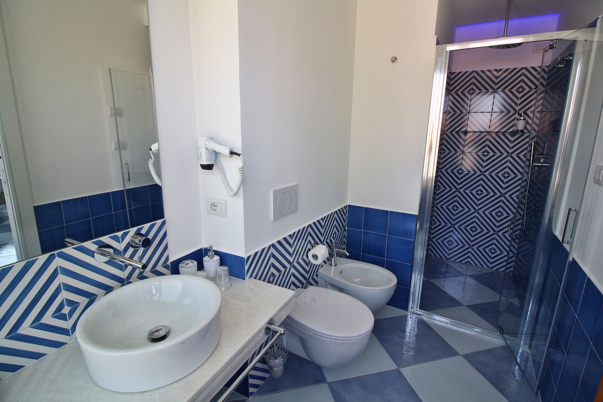 A bathroom with a white sink, two toilets, a shower, and a patterned tile backsplash.