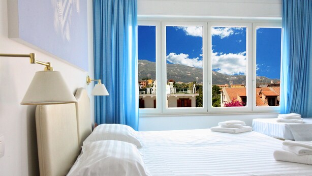 Habitación de hotel con una cama, dos lámparas de noche, una ventana que ofrece vistas a edificios y montañas, y una mesita de noche con una lámpara.