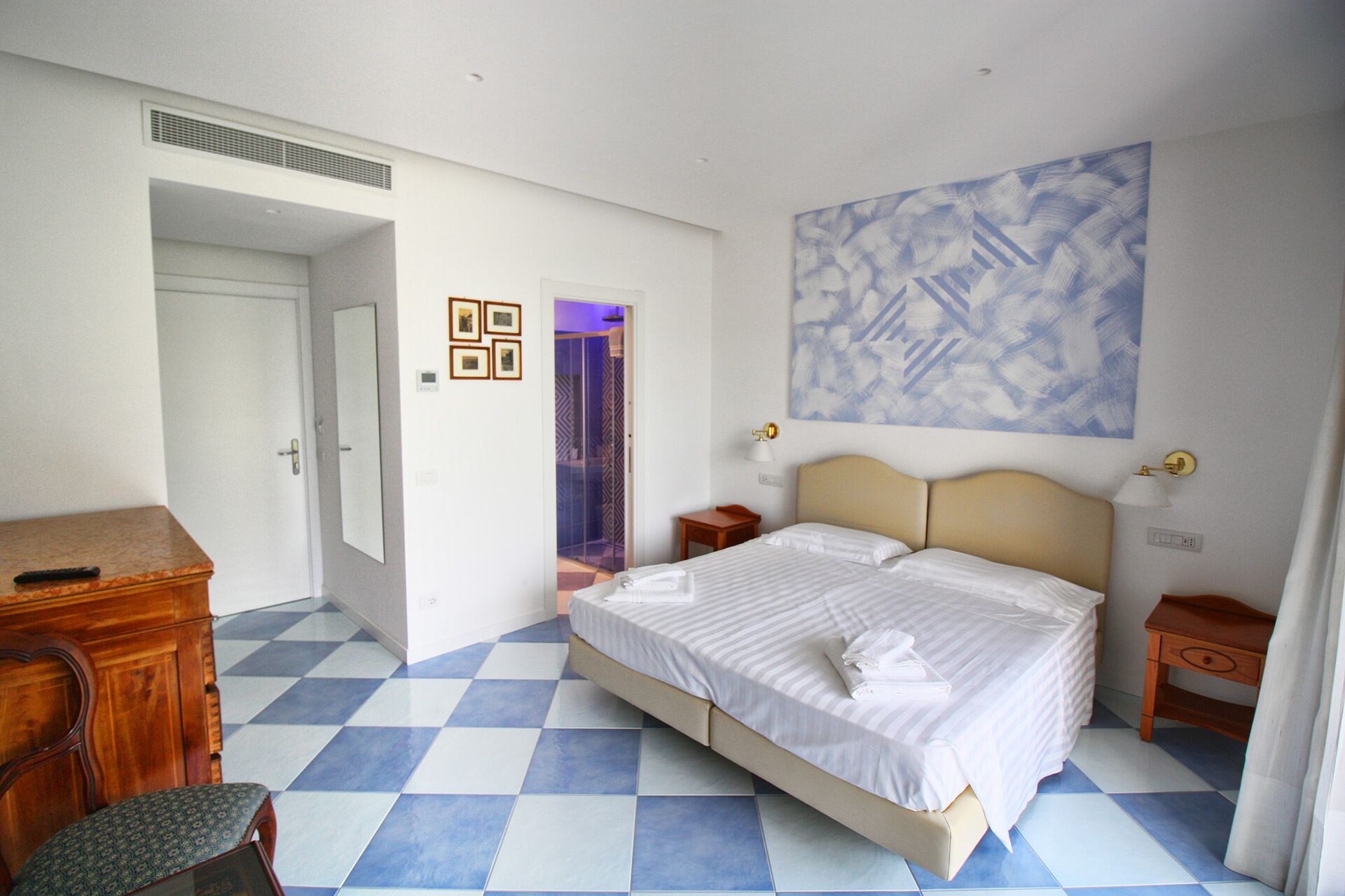 A hotel room with a checkered floor, a bed with white linens, a wooden dresser, a desk, and a wall-mounted painting.
