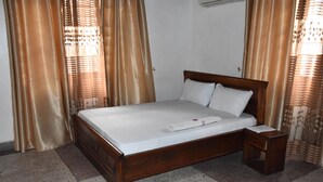 A bedroom with a wooden bed, white linens, a wooden nightstand, and beige curtains. - Dar eco backpackers (Dar es Salaam)