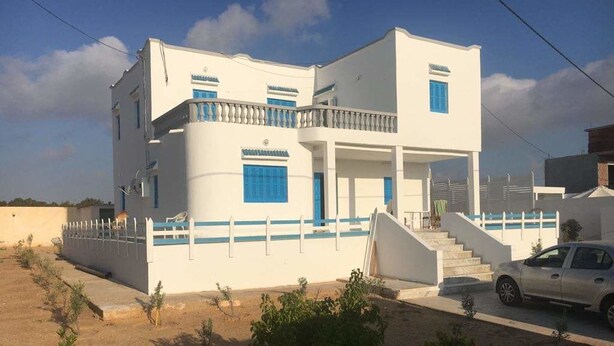 Un edificio de dos pisos con fachada blanca y detalles azules, una escalera que conduce a la entrada y un coche estacionado al frente.