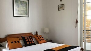 Ein ordentlich bezogenes Bett mit Holzrahmen, weißer Bettwäsche und orange-schwarzen Kissen. Darüber an der Wand hängt ein gerahmtes Bild, und daneben steht ein kleiner Tisch mit einer Lampe.