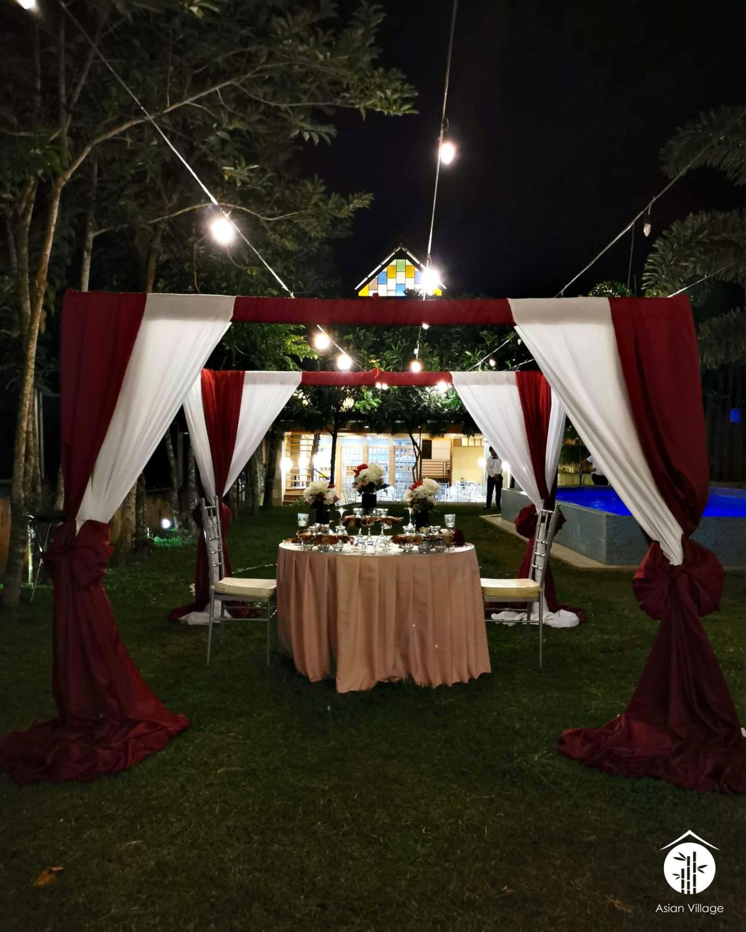 An outdoor wedding setup with a table under a canopy, surrounded by chairs and a decorative arch.