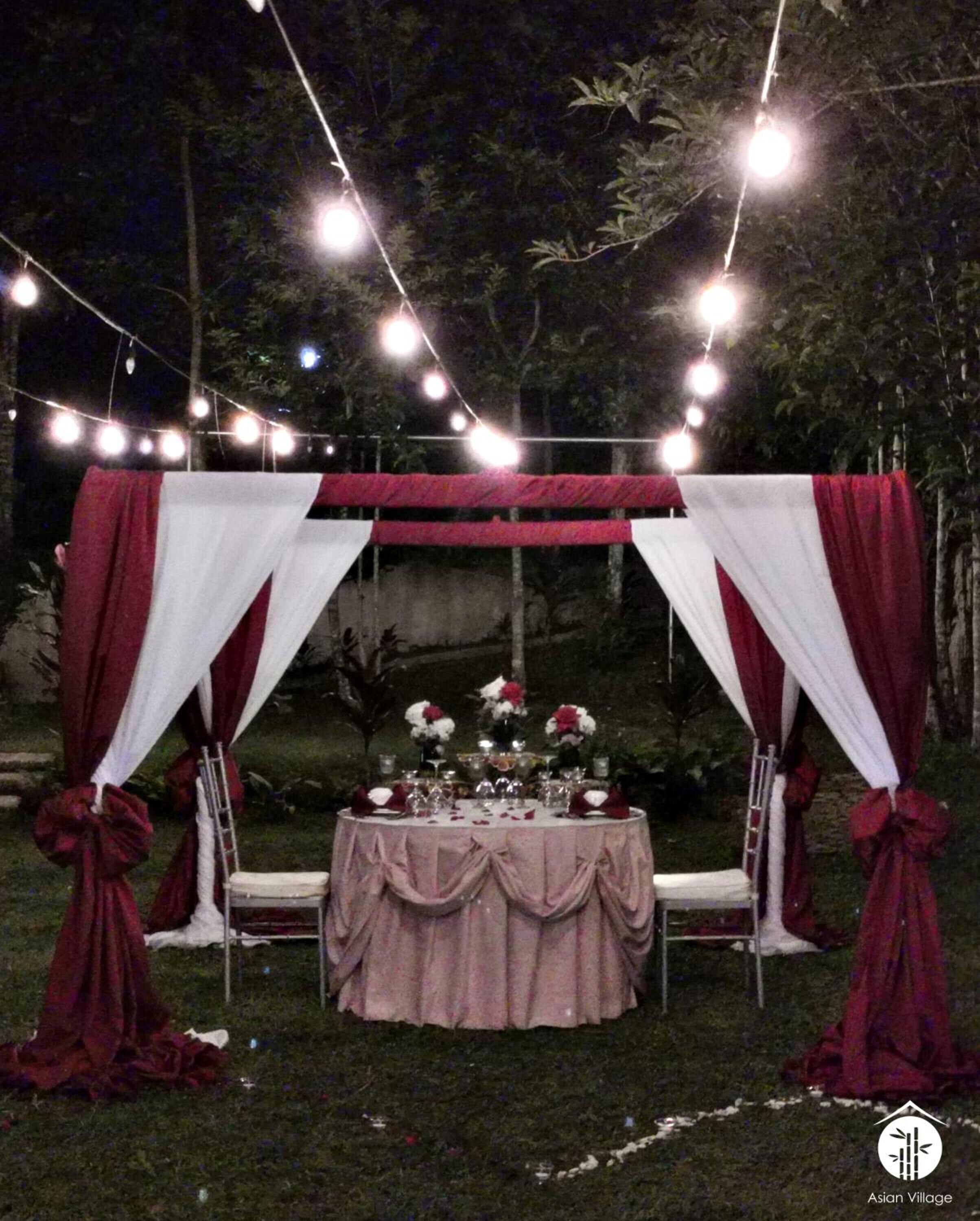 An outdoor dining setup with a table set for a meal, surrounded by draped fabric and string lights.
