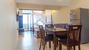 A dining area with a table, chairs, a refrigerator, and a view of a balcony with a mosquito net. - Chrystal Baharini (Diani Beach)