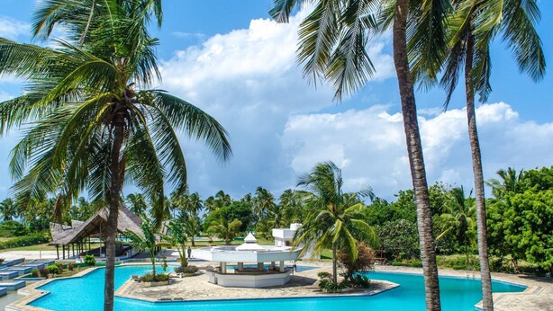 A resort pool area with a central water feature, surrounded by palm trees and a stone deck. - Chrystal Baharini (Diani Beach)
