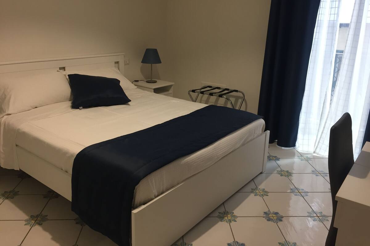 Una cama bien hecha con cubrecama y almohadas azul oscuro, una mesita de noche con una lámpara, una silla y una ventana con cortinas.