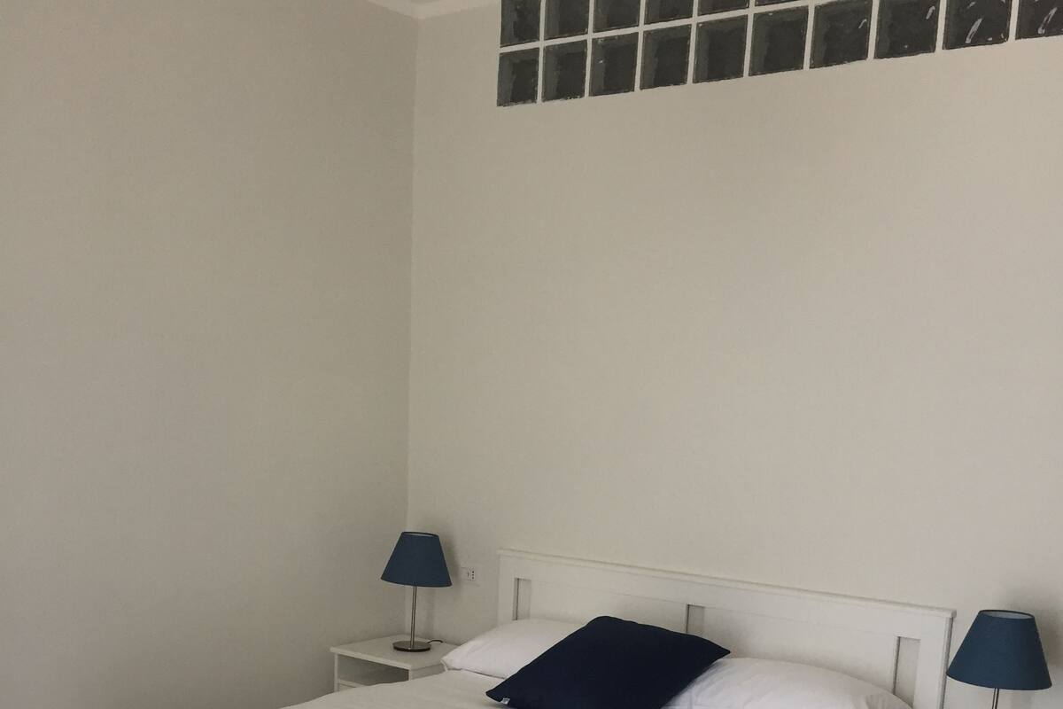 Un dormitorio con cama blanca, manta azul, dos mesitas de noche con lámparas y una ventana con diseño de cuadrícula.