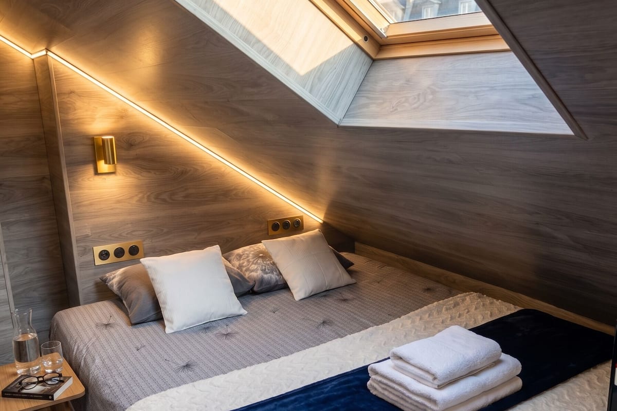 Ein modernes Schlafzimmer mit Oberlicht, einem Bett mit vielen Kissen, Nachttischen und Blick auf Gebäude.