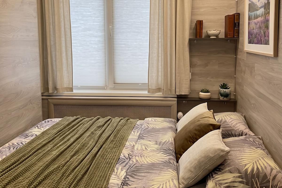 Ein Schlafzimmer mit Bett, Fenster, Vorhängen, einem Regal mit Büchern und einer Pflanze sowie einem gerahmten Bild an der Wand.