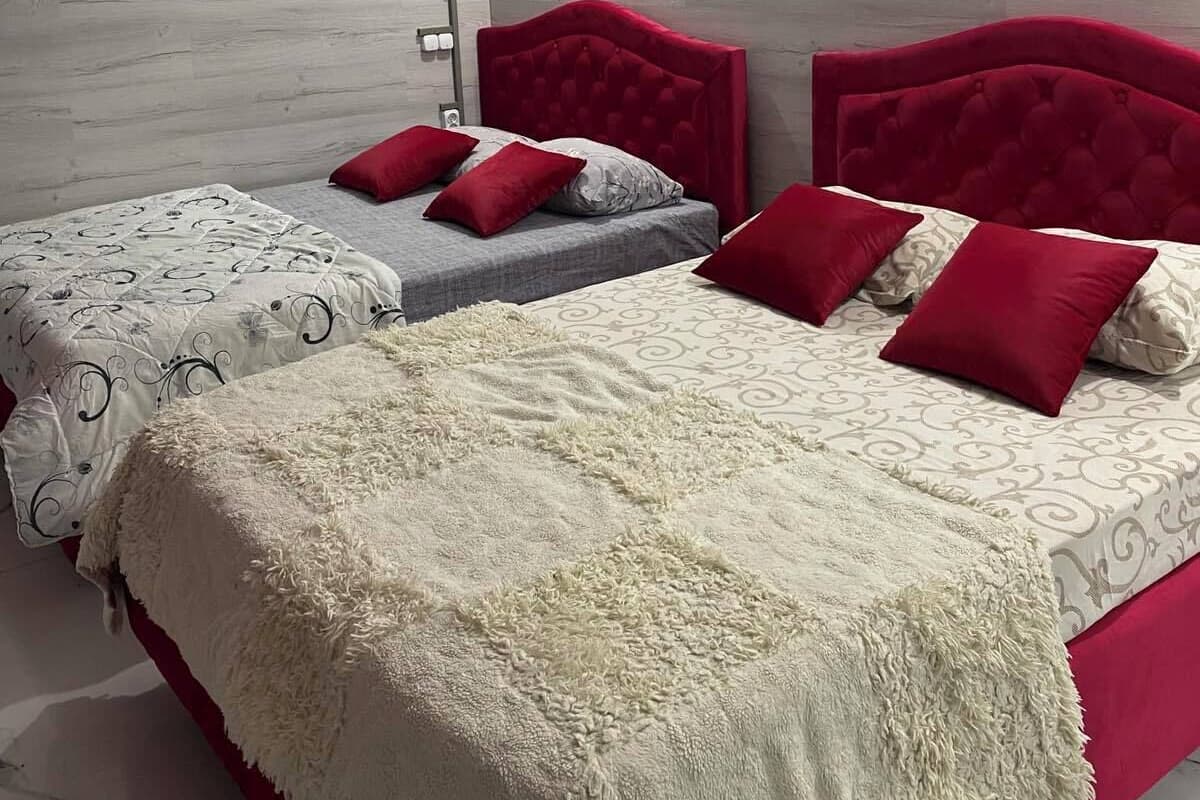 Ein Schlafzimmer mit zwei Betten, jeweils mit roten Kopfteilen und Kissen, einer weißen, flauschigen Decke und einer grauen, gemusterten Tagesdecke.