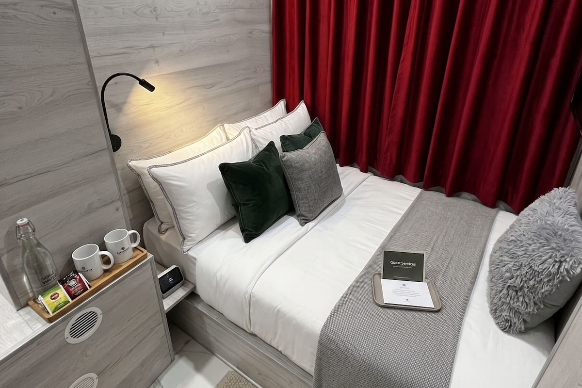 Ein modernes Hotelzimmer mit einem Holzkopfteil, einem Bett mit weißer und grauer Bettwäsche, roten Vorhängen, einem Nachttisch mit einer Lampe und einem weiteren Nachttisch mit einer Kaffeemaschine und einer Speisekarte.