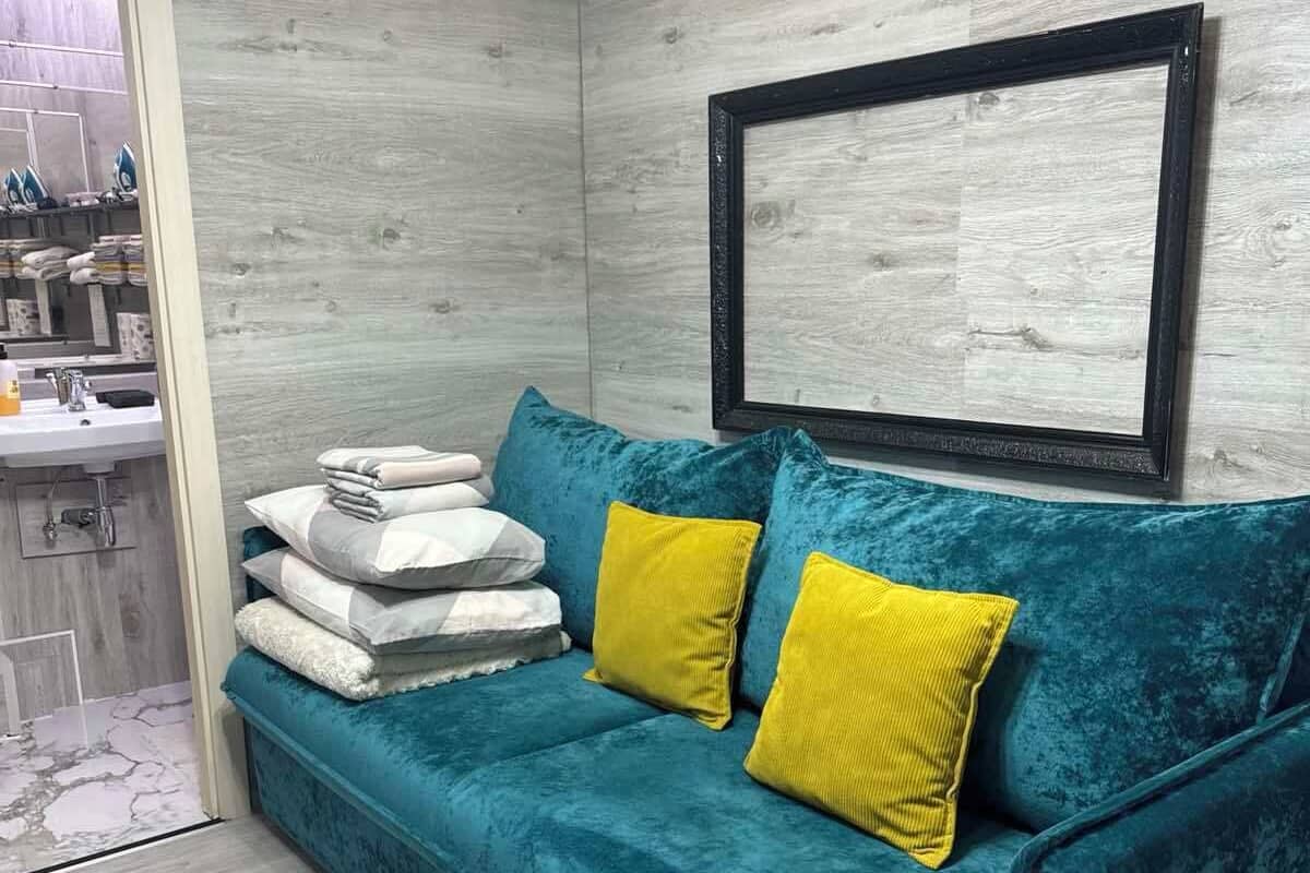 Ein smaragdgrünes Samtsofa mit zwei gelben Kissen, eine weiße Wand mit einem Bild in Schwarzrahmen und ein Holzboden.
