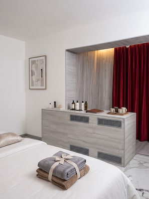 A hotel room with a bed, a vanity with toiletries, a framed picture on the wall, and a red curtain.