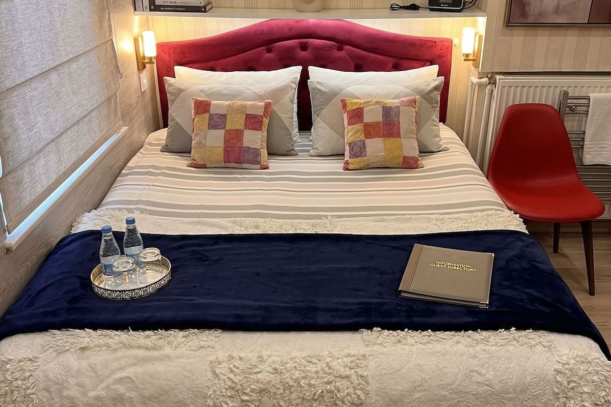 Ein Hotelzimmer mit einem Bett, Kissen, einem Tablett mit Flaschen, einem Buch und einem roten Sessel.