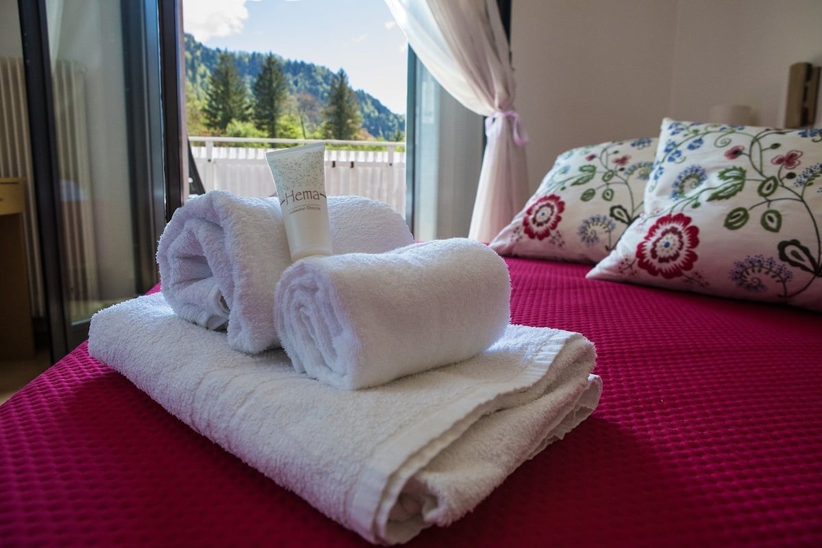 Een netjes opgemaakt bed met een roze sprei, een opgerolde witte handdoek en een fles Hema shampoo.