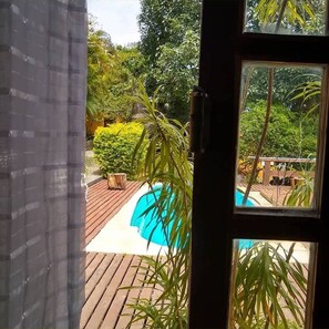 A view through a window of a wooden deck with a swimming pool, surrounded by lush greenery. - Mira Playa Geribá (Búzios)