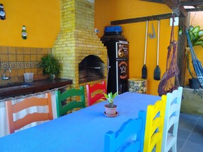A dining area with a brick fireplace, a small table with a blue tablecloth, and colorful chairs. - Mira Playa Geribá (Búzios)