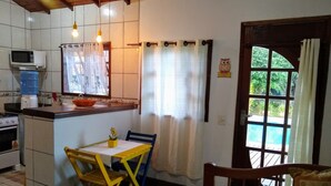 A compact kitchen with a microwave, sink, and stove. There is a dining table with chairs and a small potted plant. A window with curtains is visible, and a door leads to an outdoor area with a pool. - Mira Playa Geribá (Búzios)
