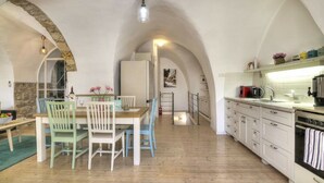Een ruime keuken met witte kastjes, een houten eettafel en stoelen. Een stenen boogpoort leidt naar een andere kamer.