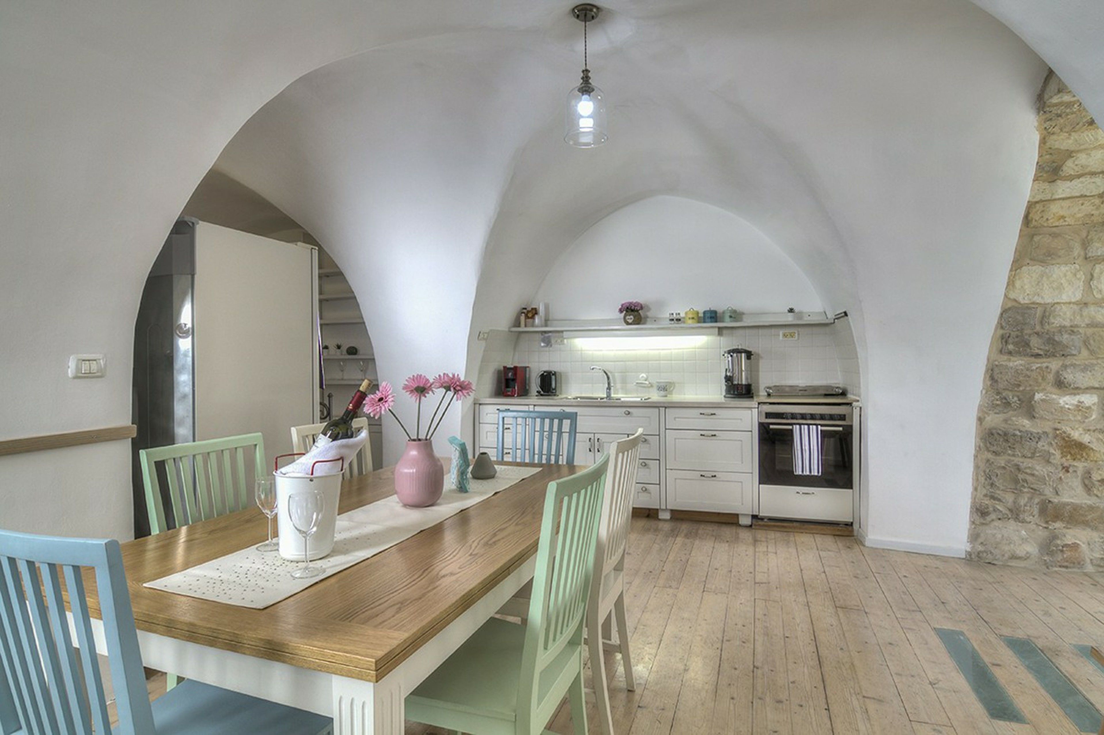 Un comedor con una mesa de madera, sillas y un florero con flores. Se observa una cocina al fondo con armarios blancos y una pared de piedra.