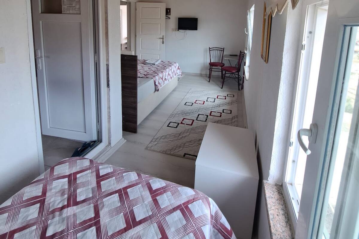 Ein Schlafzimmer mit einem Bett, einem Schreibtisch und einem Stuhl. Eine Tür führt in einen anderen Raum, und ein Fenster ist mit Vorhängen versehen.