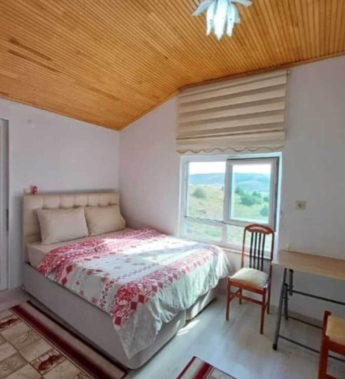 Een slaapkamer met een bed, een stoel, een bureau, een raam met uitzicht en een houten plafond.