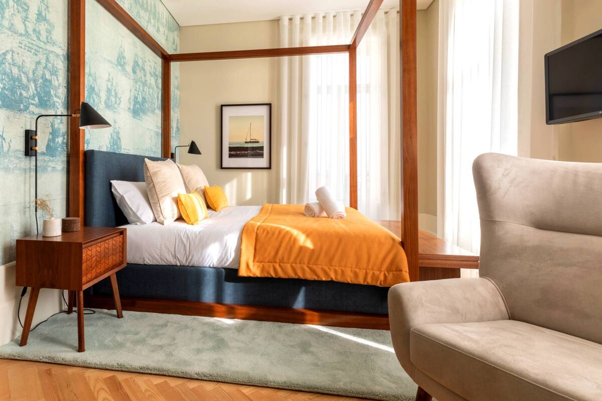 A four-poster bed with a blue headboard, a beige sofa, a wooden nightstand, and a framed picture on the wall.