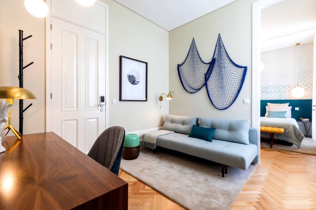 A modern living room with a wooden dining table, a grey sofa with blue cushions, a patterned rug, and a wall-mounted hammock.