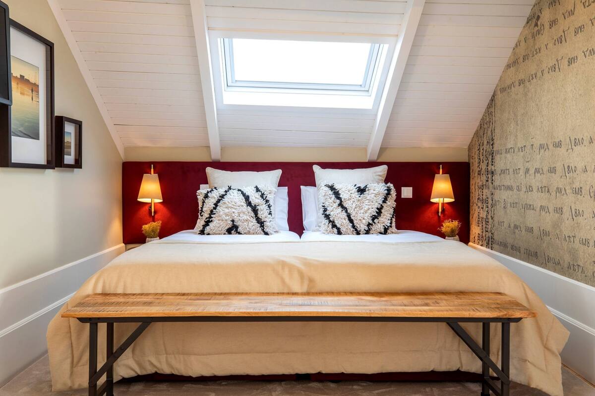 A bedroom with a skylight, a bed with patterned pillows, two bedside lamps, and a wooden bench.