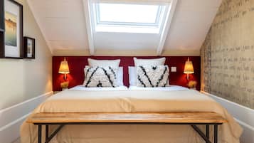 Quarto com claraboia, cama com almofadas estampadas, duas luminárias de cabeceira e um banco de madeira.