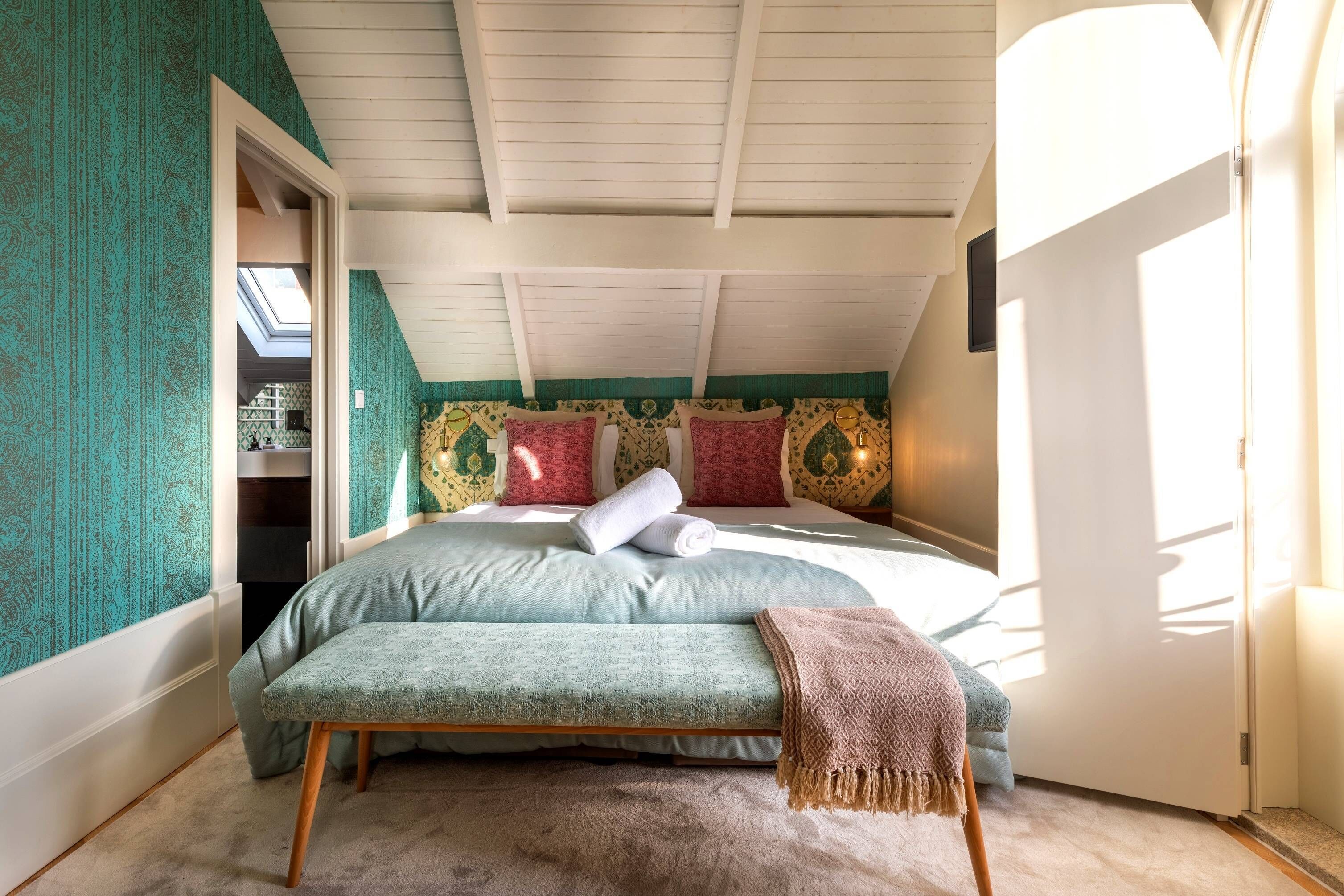 Une chambre avec un puits de lumière, un lit recouvert d’une couverture verte, un banc et des coussins à motifs.
