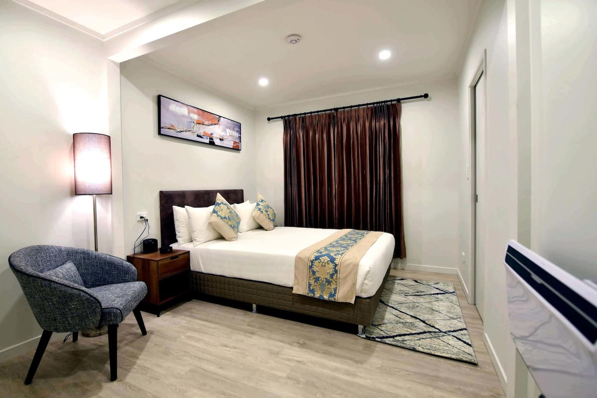 Quarto de hotel com uma cama, uma cadeira, uma mesa de cabeceira, um abajur, um quadro e uma janela com cortinas.