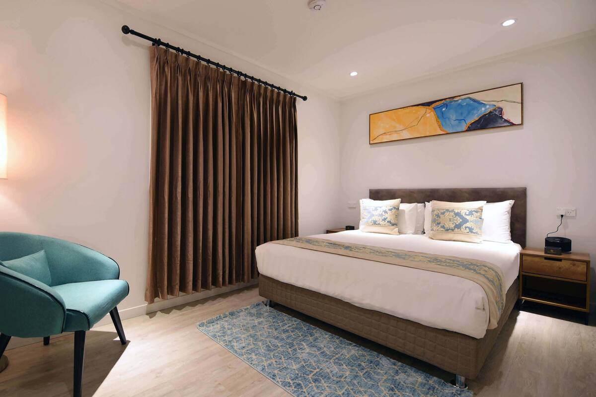 Quarto de hotel com cama, poltrona, luminária, pintura e janela com cortinas.