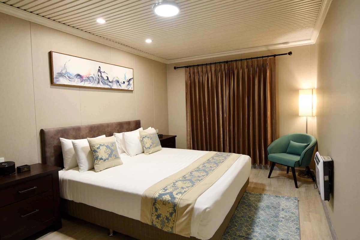 Quarto de hotel com uma cama grande, mesa de cabeceira, escrivaninha, cadeira, abajur e um quadro na parede.